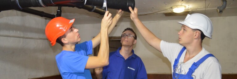 Drei Personen in Arbeitskleidung arbeiten zusammen an einer Rohrinstallation in einem Keller.