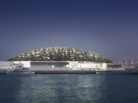 A modern architectural structure with a domed roof is illuminated at dusk, surrounded by water and city skyline in the background.