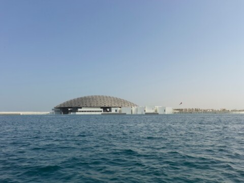 A large modern architectural structure with a dome, viewed from water under a clear blue sky.