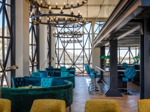 A modern interior featuring green and blue velvet seating, large chandeliers, and large windows with geometric patterns.