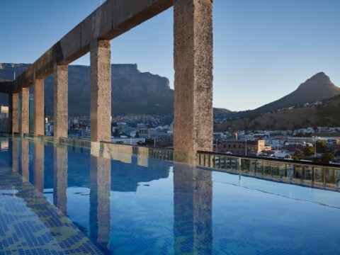 A scenic view of a swimming pool reflecting the sky and mountains, framed by large, modern stone arches.