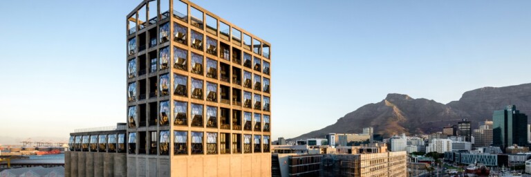 A modern building with a distinct geometric design, featuring large windows and set against a mountain backdrop.