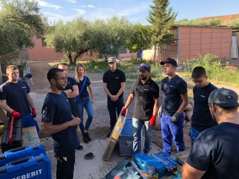 Eine Gruppe von Personen, die in Arbeitskleidung versammelt sind und einer Person zuhören, die etwas erklärt. Im Hintergrund sind Bäume und ein Gebäude sichtbar.