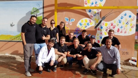 Eine Gruppe von Personen posiert vor einer bunten Wandmalerei, die einen Schmetterling zeigt, der aus Handabdrücken besteht.