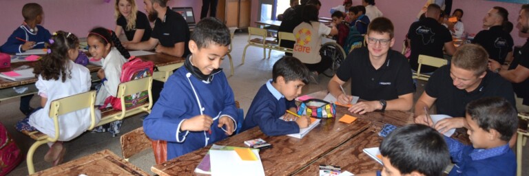 Eine Gruppe von Schülern und Freiwilligen, die in einem Klassenzimmer an verschiedenen Aktivitäten arbeiten, umgeben von bunten Wandgestaltungen.