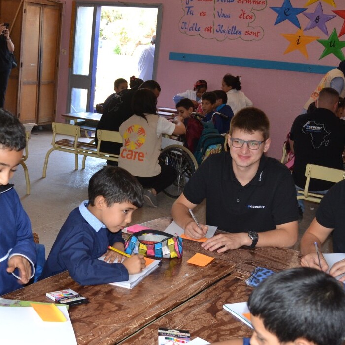 Eine Gruppe von Schülern und Freiwilligen, die in einem Klassenzimmer an verschiedenen Aktivitäten arbeiten, umgeben von bunten Wandgestaltungen.