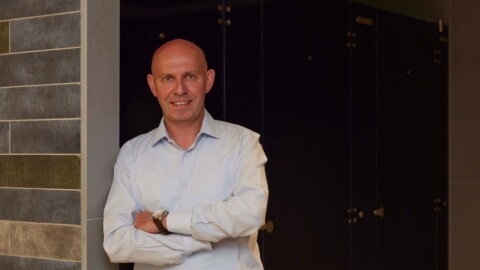 A smiling man with blue shirt standing with arms crossed in front of a door.