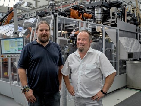 Two men stand in front of a large industrial machine with robotic arms and control screens in a factory setting.