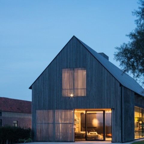A modern wooden house with large windows, set against a twilight sky and surrounded by greenery.