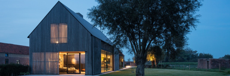 Ein modernes, zweigeschossiges Holzhaus mit grossen Fenstern, umgeben von einem grünen Rasen und einem Baum, bei Abenddämmerung.