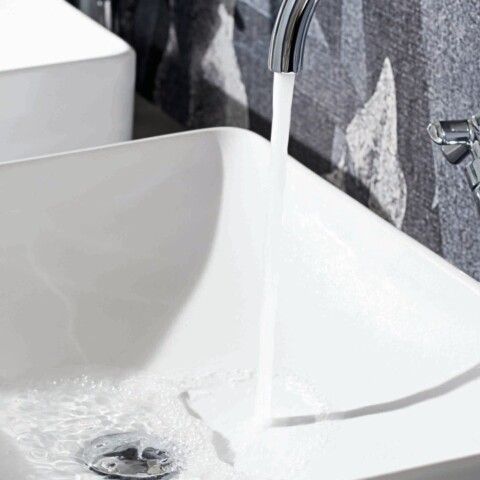 Zwei moderne, weisse Waschbecken vor einer Wand, eines mit fliessendem Wasser.