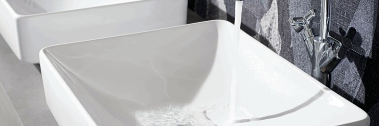 Zwei moderne, weisse Waschbecken vor einer Wand, eines mit fliessendem Wasser.