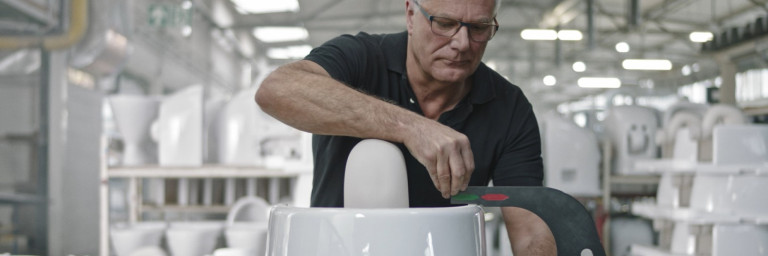 Ein älterer Mann mit Brille arbeitet in einer Fabrik an einer grossen weissen Keramikschale und misst sie mit einem Werkzeug.