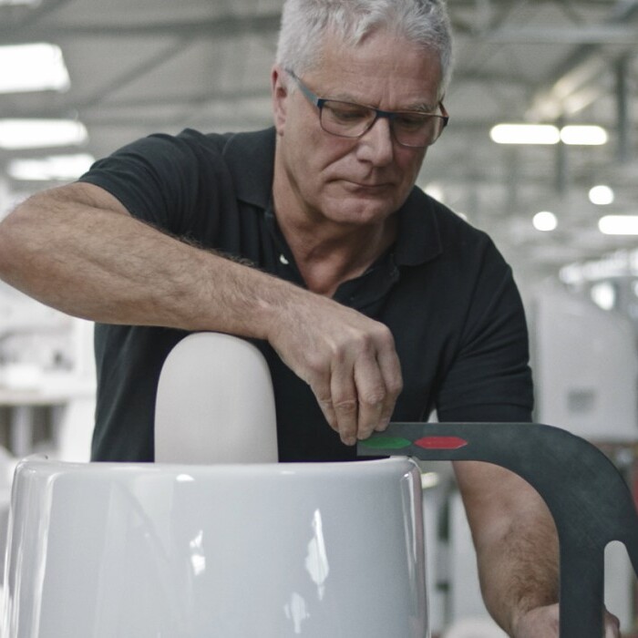 Ein älterer Mann mit Brille arbeitet in einer Fabrik an einer grossen weissen Keramikschale und misst sie mit einem Werkzeug.