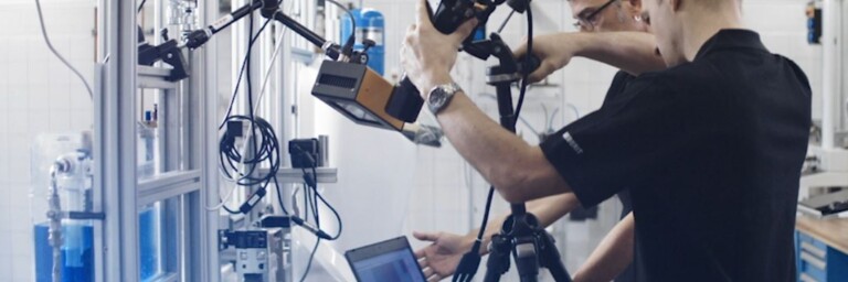 Two technicians working in a laboratory setting, one setting up a tripod while the other has a laptop next to various scientific equipment.