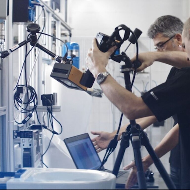 Two technicians working in a laboratory setting, one setting up a tripod while the other has a laptop next to various scientific equipment.