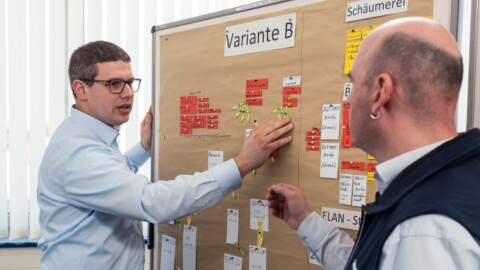 Two individuals engage in a discussion, pointing at a board filled with sticky notes.
