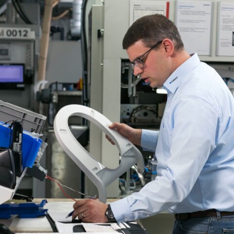 A man in a light blue shirt is focused on inspecting a component, while another person is visible in the background.