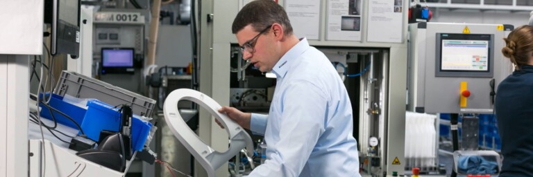 A man in a light blue shirt is focused on inspecting a component, while another person is visible in the background.