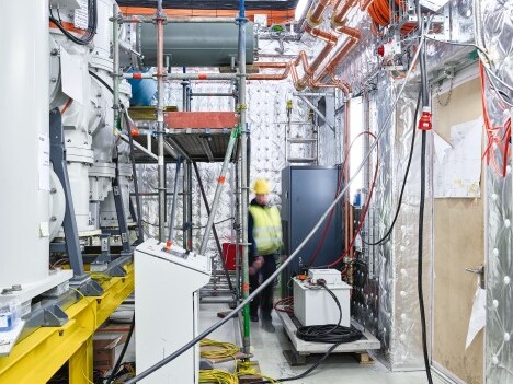 Arbeiter in Sicherheitskleidung in einem technisch ausgestatteten Arbeitsumfeld mit Maschinen, Rohren und Kabeln.