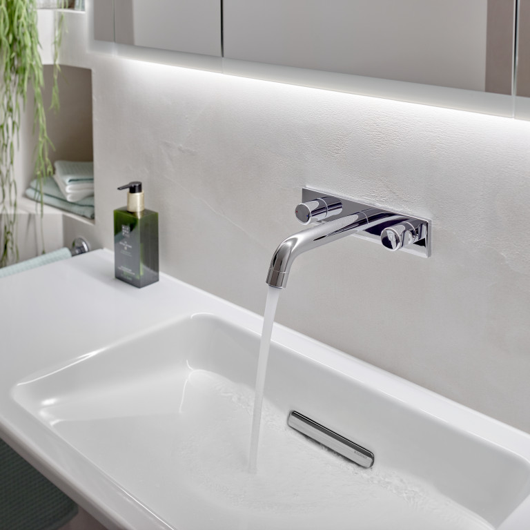 A modern bathroom sink with a chrome faucet, water flowing into a white basin, and green plant decor in the background.