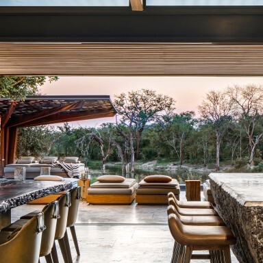 Moderner Essbereich mit Tageslicht, einem grossem Holztisch, stilvollen Sitzgelegenheiten und Blick auf die üppige Vegetation draussen.