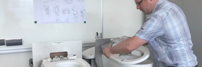 A person working on a toilet installation, with two toilets visible and technical drawings on the wall.