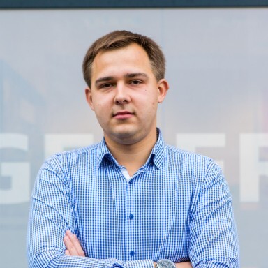 A young man stands in front of a translucent backdrop featuring the word "GEBRIT." He wears a blue checked shirt.