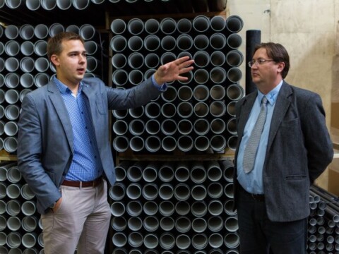 Two men stand in a warehouse filled with stacked cylindrical pipes; one gesturing while speaking, the other listening.