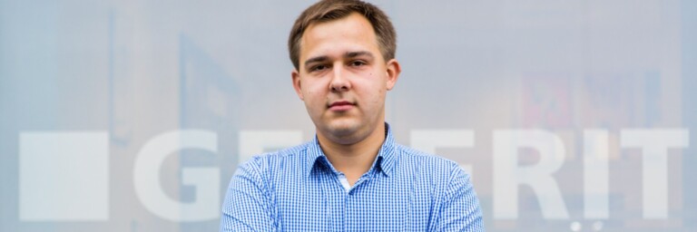 A young man stands in front of a translucent backdrop featuring the word "GEBRIT." He wears a blue checked shirt.