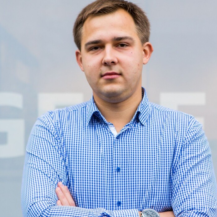 A young man stands in front of a translucent backdrop featuring the word "GEBRIT." He wears a blue checked shirt.