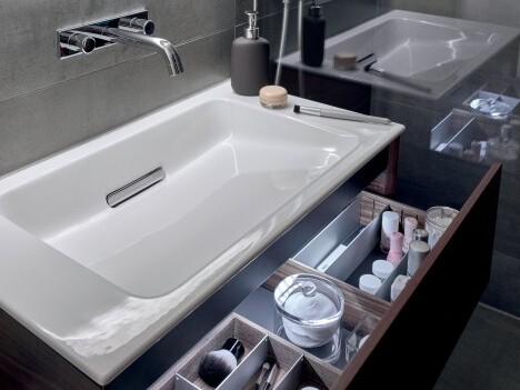 Modernes weisses Waschbecken, eine Wandarmatur und offene Schubladen mit Kosmetikprodukten.