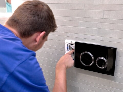 Ein Handwerker installiert eine schwarze Betätigungsplatte für eine Toilette an einer Wand aus grauen Fliesen.