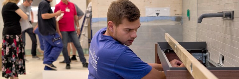 Ein Handwerker in blauer Kleidung installiert ein Waschbecken, während andere Personen im Hintergrund diskutieren.