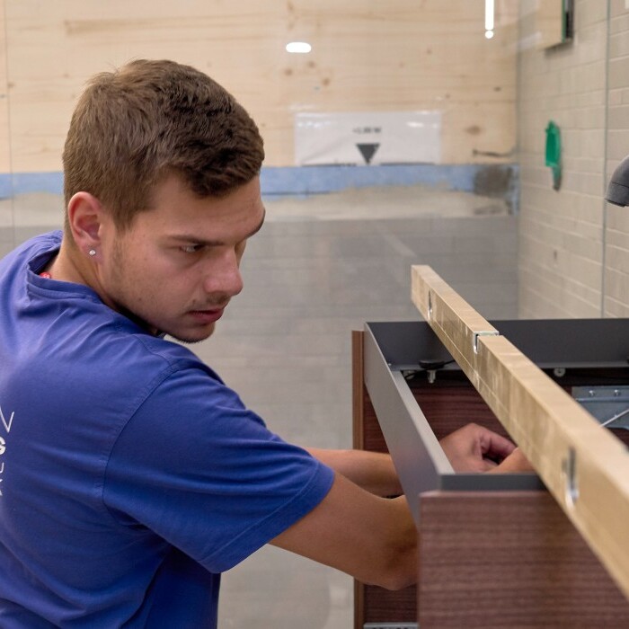 Ein Handwerker in blauer Kleidung installiert ein Waschbecken, während andere Personen im Hintergrund diskutieren.