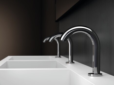 Three modern chrome faucets above a white countertop with basins, set against a dark wall.
