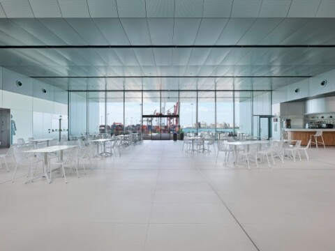 Modern cafeteria with white tables and chairs, large windows, and a kitchen area in the background.