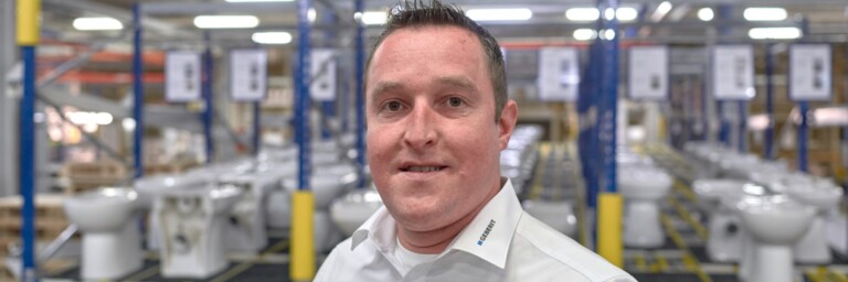 Ein Mann in einem hellen Hemd steht in einer Fabrikhalle mit Toiletten im Hintergrund.