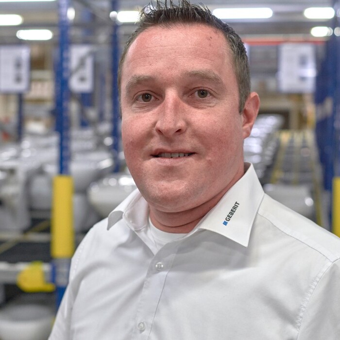 Ein Mann in einem hellen Hemd steht in einer Fabrikhalle mit Toiletten im Hintergrund.