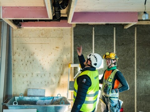 Zwei Arbeiter auf einer Baustelle, einer zeigt auf die Decke, während der andere aufmerksam zuhört.