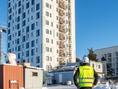 Ein Bauarbeiter in Warnweste betrachtet einen mehrstöckigen Gebäudekomplex auf einer Baustelle im Winter.