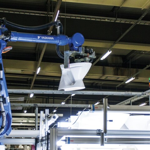Ein blauer Industrieroboterarm hebt eine weisse Toiletten Keramik in einer Fabrikumgebung.