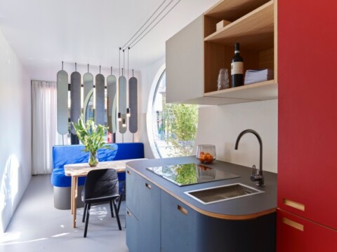 Moderne Küche mit grauem Arbeitsbereich, rotem Schrank und angrenzendem Essbereich mit blauem Sofa und Holztisch.