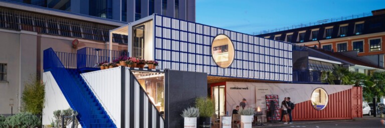 Ein modernes Gebäude, das aus mehreren Container-Einheiten besteht, mit einer blauen Treppe und Pflanzen im Vordergrund, bei Abenddämmerung.