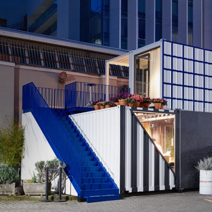 Ein modernes Gebäude, das aus mehreren Container-Einheiten besteht, mit einer blauen Treppe und Pflanzen im Vordergrund, bei Abenddämmerung.