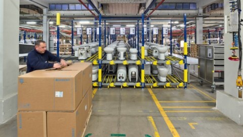 A warehouse setting with a worker handling a large cardboard box in front of storage racks filled with toilet fixtures.