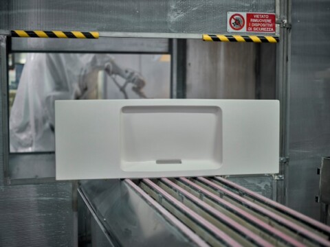 Eine weisse Waschbeckenkeramik auf einem Förderband in einer industriellen Umgebung mit Sicherheitszeichen im Hintergrund.