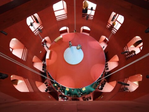 A circular indoor space with red walls and multiple levels, featuring people in the building.