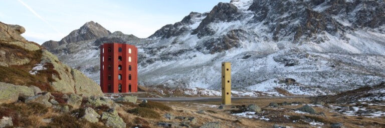 Two unique, colorful towers, one red and one yellow, situated in a mountainous landscape with patches of snow.