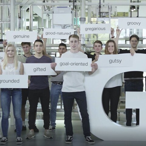 Eine Gruppe junger Menschen steht zusammen und hält Schilder mit positiven Adjektiven, während sie vor einem grossen Buchstaben "G" posieren.